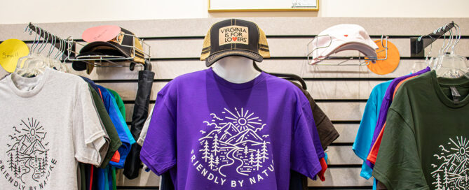 A wall display featuring a manikin wearing a purple Friendly by Nature Harrisonburg t-shirt and a Virginia is for Lovers trucker hat.