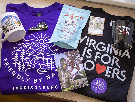 Items sold in the Rocktown Gift Shoppe including a Harrisonburg mug, a Friendly by nature t-shirt, a Virginia is for Lovers t-shirt, Redrocker Peppermint candy, a spring postcard of the Harrisonburg court house, a purple Friendly by Nature magnet and a Shenandoah National Park magnet.