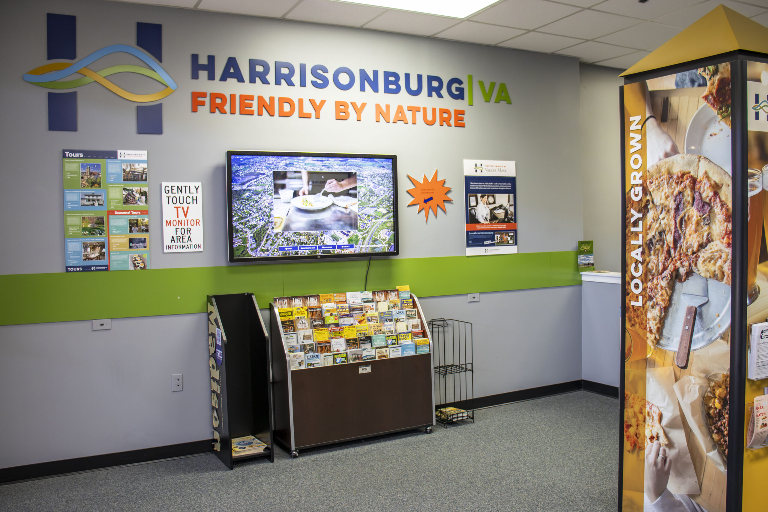 Wall with brochures and interactive T-V under Harrisonburg V-A Friendly by Nature logo at the Visitor Center in the Valley Mall.