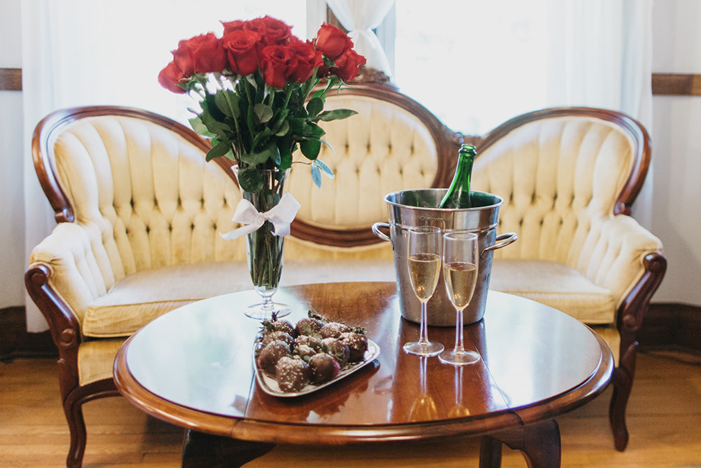 A vase of roses, champagne, and chocolate strawberries on a table in front of a couch