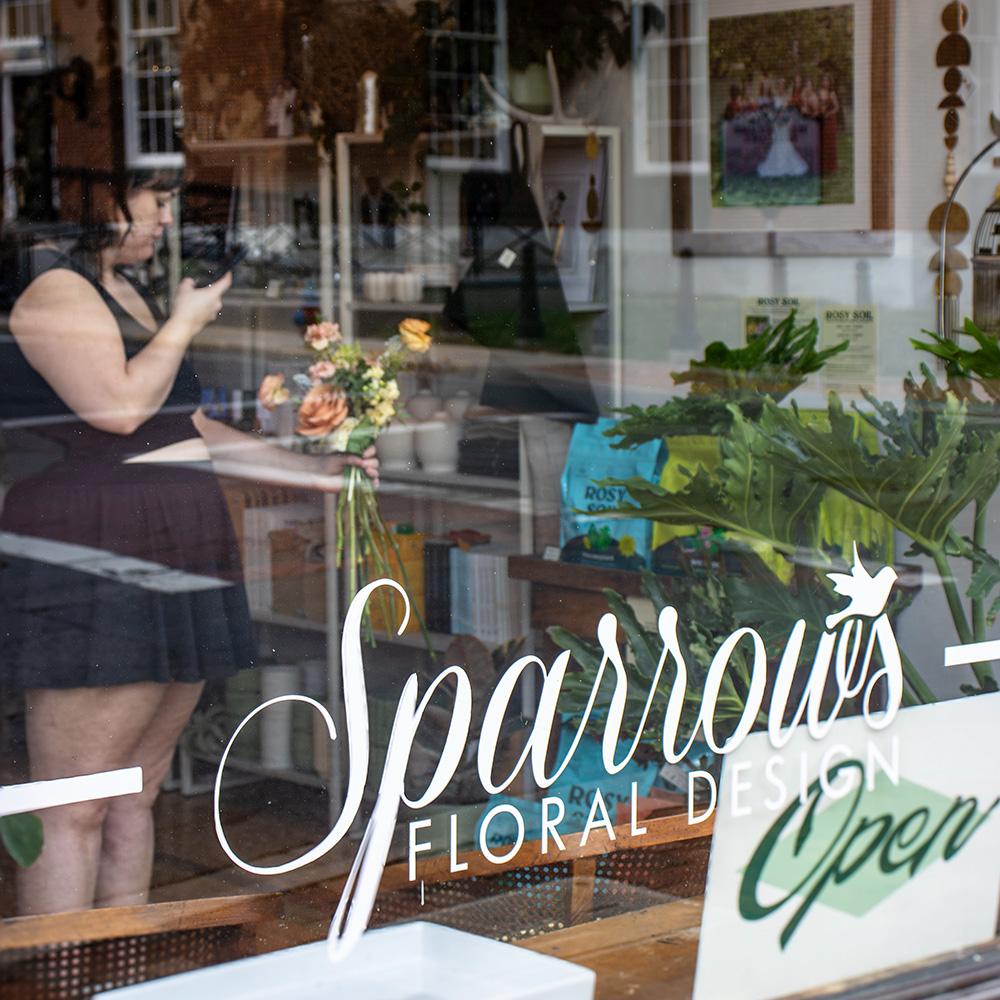 Women in a store window of Sparrows Floral Design taking a photo of a bouquet of flowers