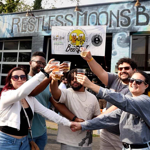 A group of people raising their beer glasses to cheer together in front of Restless Moons Brewing
